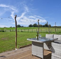 A garden with a wooden deck a gray wicker table and sofa with a wine bottle and glasses at Maple in Wootton Fitzpaine near Charmouth