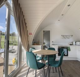 A kitchen and dining area with a round wooden table and teal chairs a toaster and oven in a white interior at Maple in Wootton Fitzpaine near Charmouth