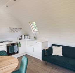 A kitchen with white cabinets and appliances next to a dark green sofa with cushions at Maple in Wootton Fitzpaine near Charmouth