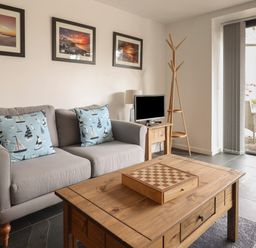 A living room with a gray sofa and blue cushions with sailboat patterns a wooden coffee table with a chessboard a small wooden side table with a lamp and a TV a wooden coat rack and a glass door leading outside at The Hideaway in Uplyme near Lyme Regis