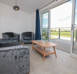A living room with a sofa, two armchairs, a wooden coffee table with flowers, and large sliding glass doors opening to a deck at Buttercup Lodge in Norton near Dartmouth