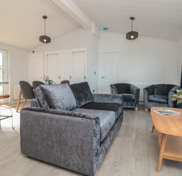 A living room with a grey sofa two armchairs a wooden coffee table with flowers and magazines and a dining table with chairs at Buttercup Lodge Norton near Dartmouth