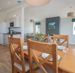 A dining area with a wooden table and chairs set with plates and glasses next to a kitchen with white cabinets and a white refrigerator at Honeysuckle Lodge in Norton near Dartmouth