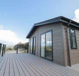A wooden deck with glass railing attached to a small building with sliding glass doors at Primrose Lodge in Norton near Dartmouth