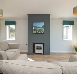 A living room with two beige sofas a fireplace with a white stove a painting above it two small windows with green blinds and flowers in the corner at Primrose Lodge in Norton near Dartmouth