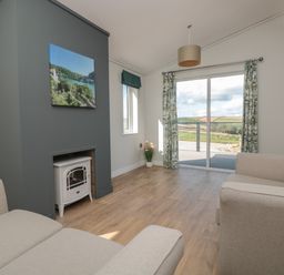 A living room with two beige sofas a white stove in a grey fireplace and sliding glass doors with curtains at Primrose Lodge in Norton near Dartmouth