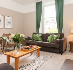 A living room with a brown sofa and green cushions a wooden coffee table with a vase of flowers and a tray two armchairs a floor lamp and framed pictures on a wall at Church View in Grange-Over-Sands