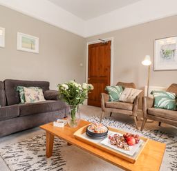 A living room with a gray sofa, two armchairs, a wooden coffee table with a vase of flowers and a tray of snacks at Church View in Grange-Over-Sands