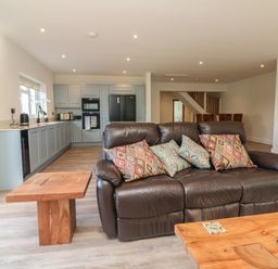 An open plan kitchen and living room with a brown leather sofa with patterned cushions a wooden coffee table and side table at Linden View in Combe Martin