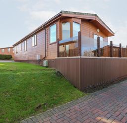 A wooden cabin with a glass railing on the deck and green grass beside a brick pathway at Serenity Sunset in Ilfracombe