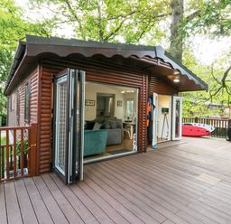 A wooden cabin exterior with an open patio door showing a living area and a wooden deck with outdoor items at Tranquil Waters in Lakeside 16 White Cross Bay near Windermere