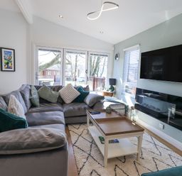 A living room with a gray sectional sofa green and white cushions a wooden coffee table a wall-mounted tv and glass doors at Tranquil Waters Lakeside 16 White Cross Bay near Windermere