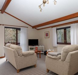 A living room with two beige sofas a TV on a stand a coffee table and windows with white curtains at Teign River Retreat in Drewsteignton near Moretonhampstead