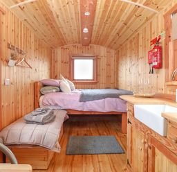 A small wooden room with a bed, bench with cushions, kitchen sink, and window at Hafan Gelli Sheperds Hut - Enlli in Pwllheli