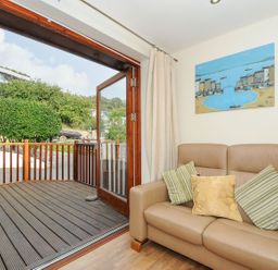 A seating area with a beige leather sofa with cushions and an open glass door leading to a wooden deck outside at Little Trevallan in Looe