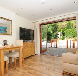 A living room with a wooden dining table and chairs a flat screen tv on a cabinet a sofa and open doors leading to a garden at Little Trevallan in Looe