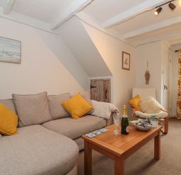 A living room with a grey sofa and yellow cushions a wooden coffee table with a bottle and glasses and a white armchair with cushions at Shell Seeker Cottage in Sewerby