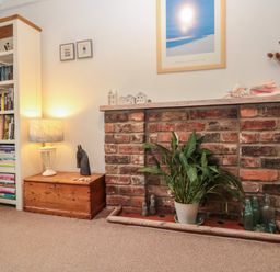 A brick fireplace with a potted plant and glass bottles next to a wooden chest with a lamp and a bookshelf in a room at Shell Seeker Cottage in Sewerby