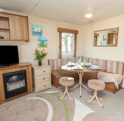 A dining area with a corner bench and stools around a table set with plates and glasses a television above a fireplace and a small drawer unit with flowers at 15 Rosewood in Great Thorness near Northwood Isle Of Wight