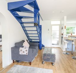 A living room and kitchen area with a blue metal staircase a gray sofa and a wooden table at Mermaid Cottage in Wakeham Portland