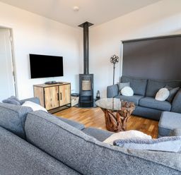 A living room with gray sofas a wooden cabinet a wall-mounted tv a wood-burning stove and a round glass coffee table at Osprey View Lodge & Hot Tub Aberfeldy