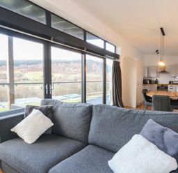 A living room with a gray sofa and cushions next to large windows showing a landscape at Osprey View Lodge & Hot Tub in Aberfeldy