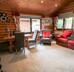 A living room with a wooden dining table and chairs a gray armchair and a red leather sofa with red cushions in a log cabin at 10 Water's Edge Morpeth near Felton