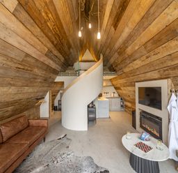 An interior view of an A-frame room with wooden walls a brown sofa a spiral staircase a kitchen a fireplace and a small table at The Tri-Pod Pantcefn near Llanarth
