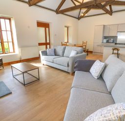 A living room with two gray sofas a coffee table a television a wood burner a dining table and a kitchen area with bar stools at The Granary in Prussia Cove near Rosudgeon