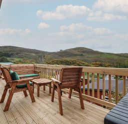 A wooden deck with outdoor furniture overlooking green hills at Y Goedlan in Tywyn