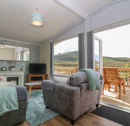A living room with armchairs blankets a rug a TV a coffee table and a balcony with wooden chairs at Y Goedlan in Tywyn
