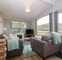 A living room with two sofas a television on a wooden cabinet a coffee table and large windows showing an outdoor view at Y Goedlan in Tywyn