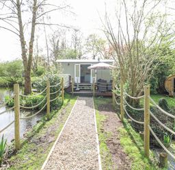A gravel pathway with rope fencing leading to a small hut with outdoor seating and an umbrella next to a pond at Damsel Shepherds Hut in Badwell Ash