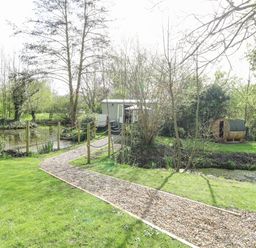 A gravel pathway leading to a small green hut and a wooden barrel sauna near a pond surrounded by trees at Damsel Shepherds Hut in Badwell Ash