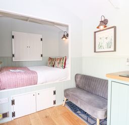 A built-in bed with colorful pillows and a pink blanket next to a gray velvet sofa in a small living area at Damsel Shepherds Hut in Badwell Ash
