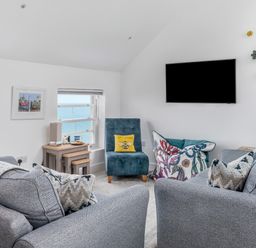 A living room with grey sofas a blue chair a wall-mounted television and windows showing the sea at Loe Cottage in Porthleven