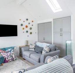 A living room with grey sofas, patterned cushions, a wall mounted TV, decorative wall art, and grey cabinets at Loe Cottage in Porthleven