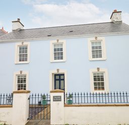 A blue two-story house with white trim and a black front door behind a blue metal fence at The Old Boathouse in Trefin near St Davids
