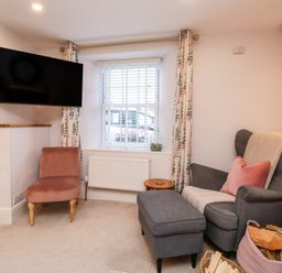 A living room with a gray armchair and ottoman a pink chair a wall-mounted television and a window with curtains at Pilchard Cottage in Dawlish