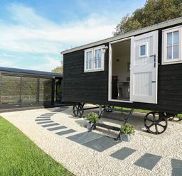 A black wooden shepherds hut with white door and windows on gravel with a stone pathway at Dragonfly Shepherd's Hut in Oakford near Aberaeron