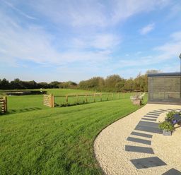 A grassy field with a fenced gate beside a path leading to a black mobile hut with stairs and potted plants at Dragonfly Shepherd's Hut Oakford near Aberaeron