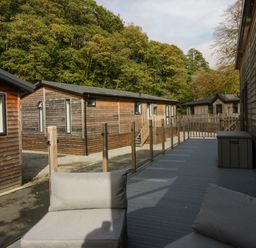 A deck with outdoor chairs and a storage box next to wooden cabins and trees in the background at The Bothy in Keswick