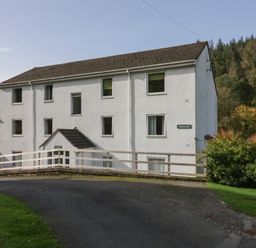 A three story white building with multiple windows and a tiled roof next to a paved road and green grass at Bobbin Mill in Keswick