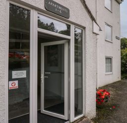 Entrance to an apartment building with a glass door and a sign reading apartments 1-6 a pot with red flowers outside Apartment 6 in Keswick