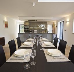A dining room with a table set with plates and glasses in front of a kitchen at The Milking Shed in Bantham