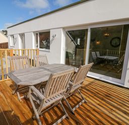 A wooden outdoor table with four chairs on a wooden deck outside a white building with sliding glass doors at Robins Downs in Ilfracombe
