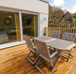 A wooden deck with a wooden table and four chairs outside a house with sliding glass doors at Robins Downs in Ilfracombe