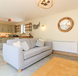 A living room with a gray sofa pillows a wooden framed circular mirror kitchen cabinets and shelves in the background at Robins Downs in Ilfracombe