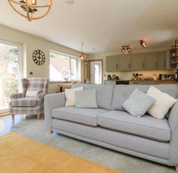A living room with a gray sofa and plaid armchair next to a sliding glass door with a kitchen in the background at Robins Downs in Ilfracombe
