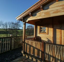A wooden cabin porch with a chair and wooden railing overlooking trees and green fields at Ballinorig Lodge in Cleator Moor